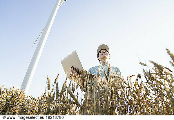 Happy farmer holding laptop standing under sky at wheat field
