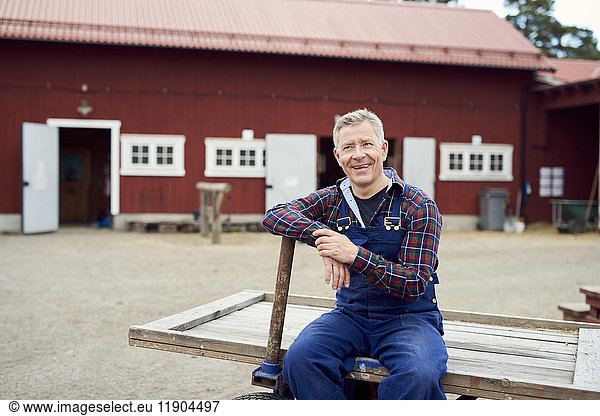 Happy farmer looking away while sitting on trailer against barn