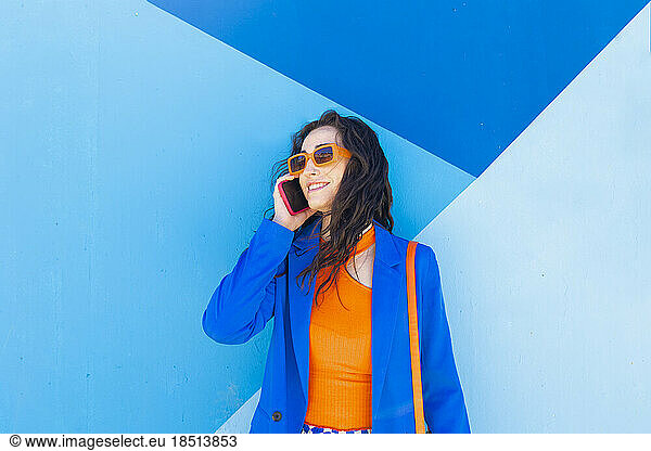 Happy fashionable woman wearing sunglasses talking on smart phone in front of blue wall