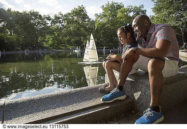 Happy father and daughter playing with remote sailboat on pond at park