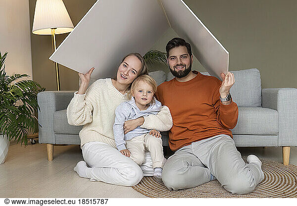 Happy father and mother with son sitting under cardboard roof at home