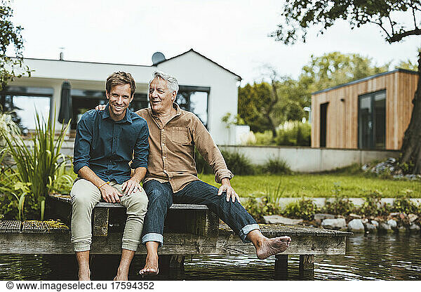Happy father and son sitting on jetty by lake at backyard