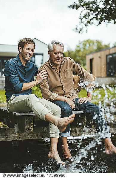 Happy father and son sitting on jetty splashing water at backyard