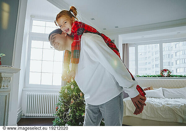Happy father giving piggyback ride to daughter near Christmas tree at home