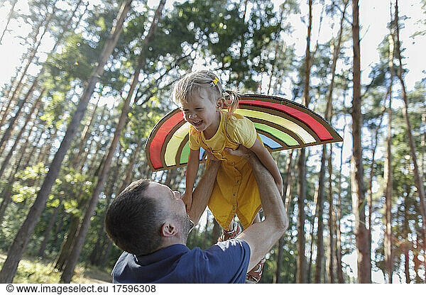Happy father lifting cheerful daughter wearing artificial wings in forest