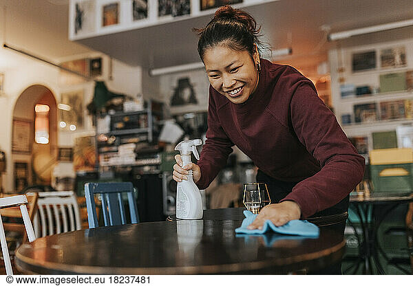 Happy female coffee shop owner with spray bottle and rag cleaning table in cafe
