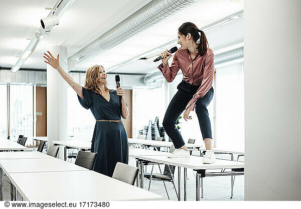 Happy female colleagues singing on microphone at conference table while taking a break in office