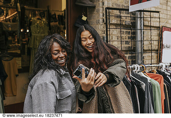 Happy female friends taking selfie through smart phone while enjoying outside clothing store