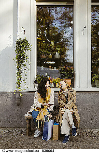 Happy female friends talking to each other while sitting on bench outside store