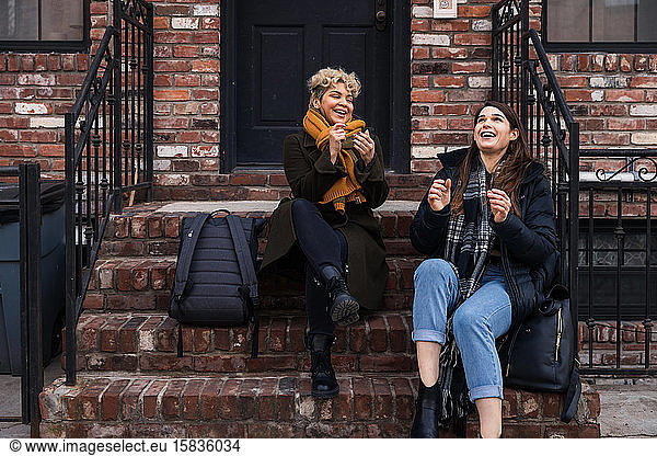 Happy female friends talking while sitting on steps in city during winter