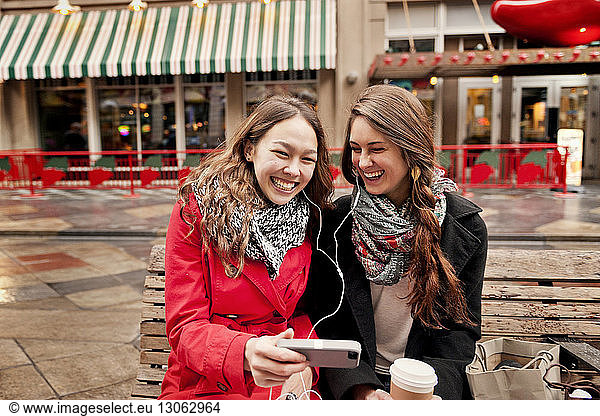 Happy female friends using phone while sitting on bench in city