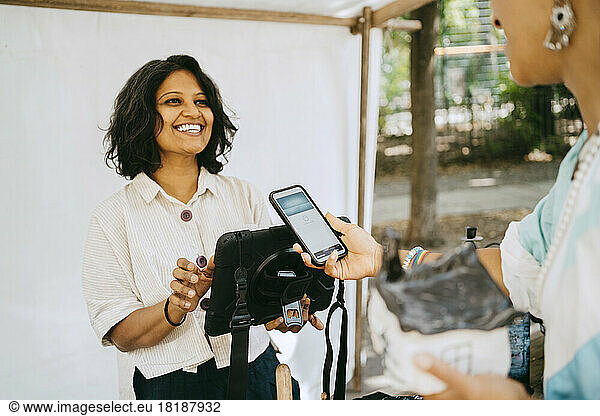 Happy female owner communicating with customer doing mobile payment at flea market