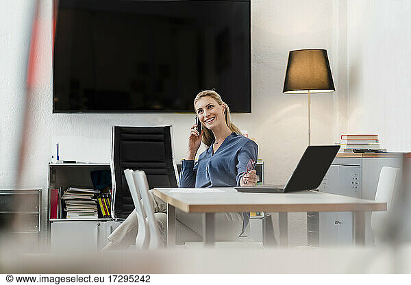 Happy female professional talking on smart phone while sitting at desk in office