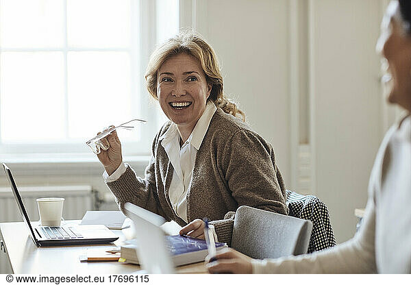 Happy female teacher holding eyeglasses looking at colleague in staffroom
