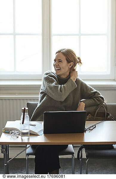 Happy female university student sitting with laptop on desk in classroom