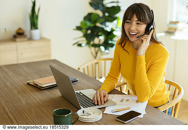 Happy freelancer talking on headset sitting with laptop at home office