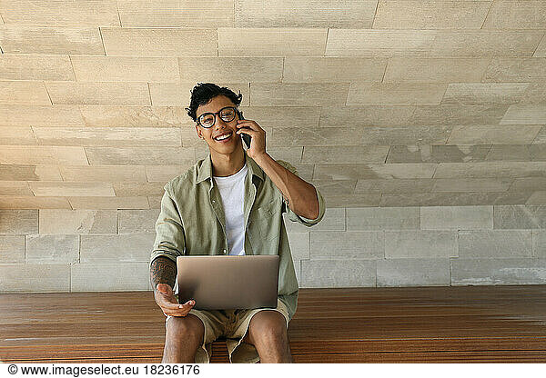 Happy freelancer talking through mobile phone sitting with laptop on bench