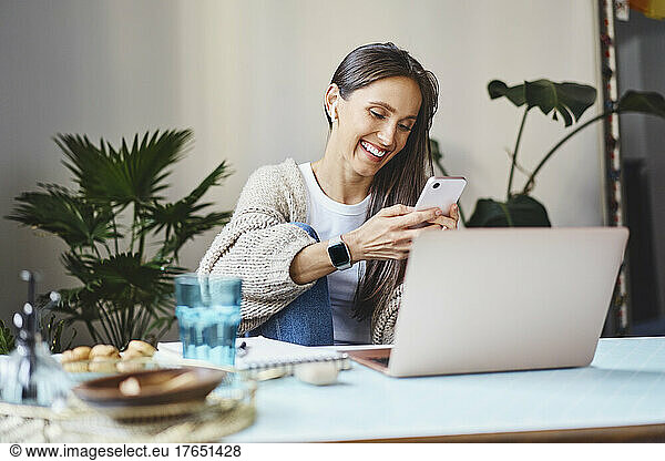 Happy freelancer using mobile phone sitting with laptop at desk