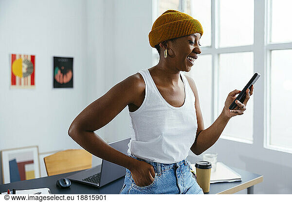 Happy freelancer using mobile phone wearing knit hat at office