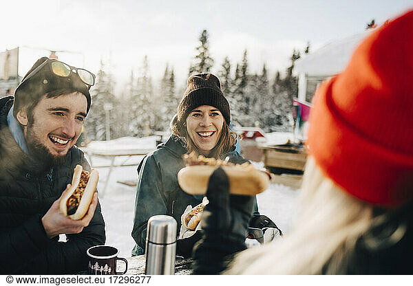 Happy friends eating hot dog while sitting at ski resort during winter