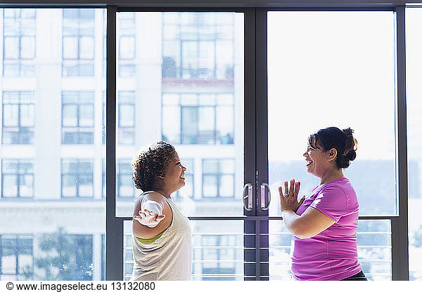 Happy friends exercising face to face while standing against window in yoga studio