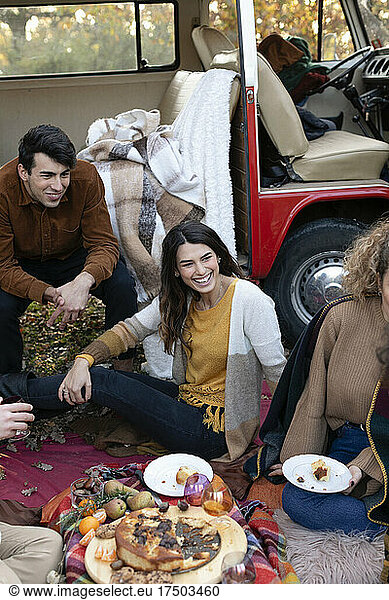 Happy friends having food on picnic outside van in autumn forest