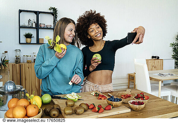 Happy friends holding fresh fruits taking selfie through mobile phone in kitchen