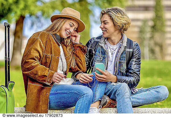 Happy friends listening music through smart phone sitting on bench