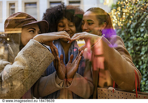 Happy friends making heart shape together with hands seen through glass