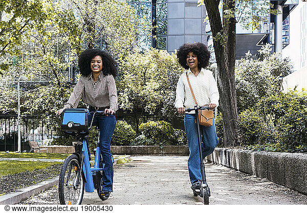 Happy friends riding electric bicycle and push scooter together in park