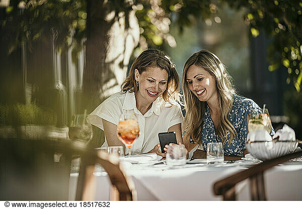 Happy friends sharing smart phone sitting at table in restaurant