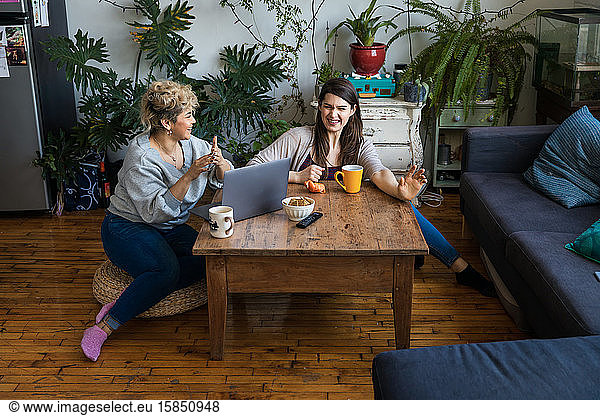 Happy friends talking while sitting with coffee mugs and laptop at table