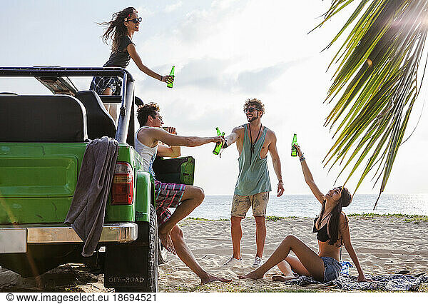 Happy friends toasting drink bottles with off-road vehicle at beach