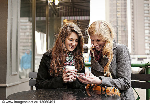 Happy friends using phone while sitting at sidewalk cafe