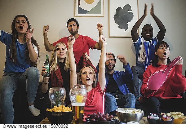 Happy friends with arms raised watching sport while sitting on sofa at home