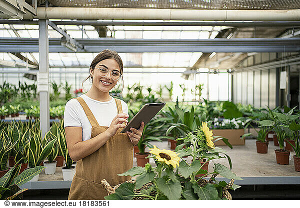 Happy gardener with tablet PC standing in plant nursery