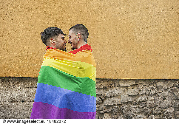Happy gay couple covered in rainbow flag by wall