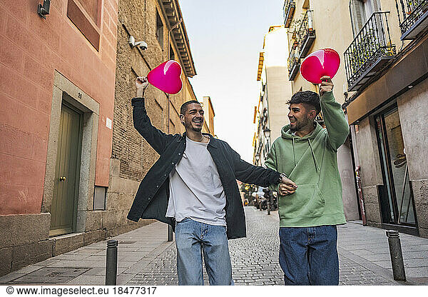 Happy gay couple having fun with red heart shape balloons amidst buildings at street