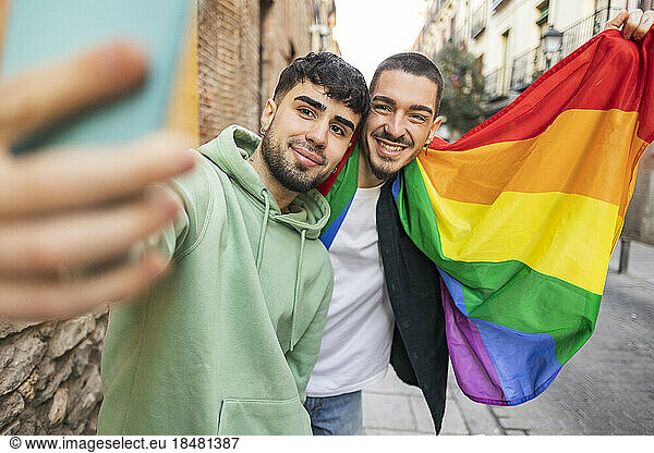 Happy gay couple with rainbow flag taking selfie through smart phone on footpath