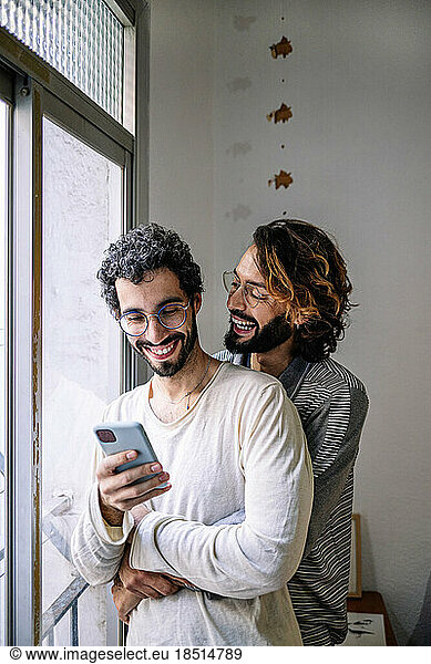 Happy gay man embracing boyfriend using smart phone near window at home