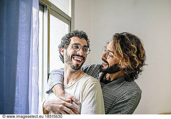 Happy gay man with arm around boyfriend near window at home