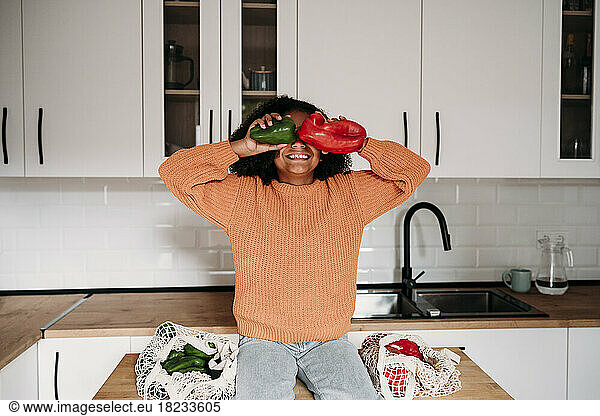 Happy girl covering eyes with bell peppers sitting in kitchen at home