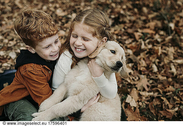 Happy girl embracing golden retriever puppy by brother on autumn leaves in forest