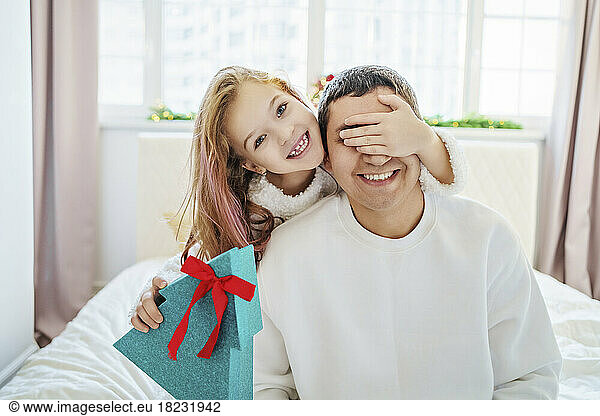 Happy girl holding Christmas gift and covering eyes of father sitting on bed at home