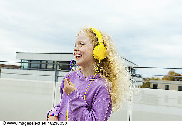 Happy girl holding small flower listening music through headphones at rooftop
