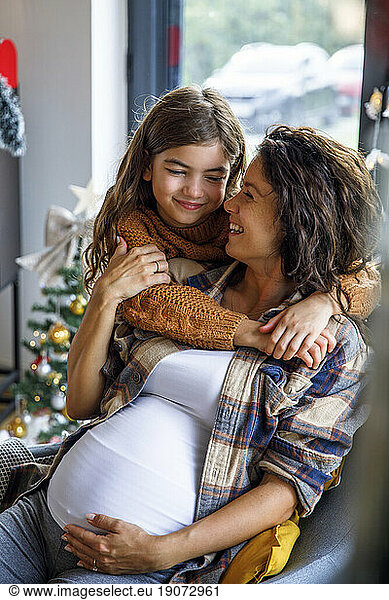 Happy girl hugging expectant mother sitting on chair at home