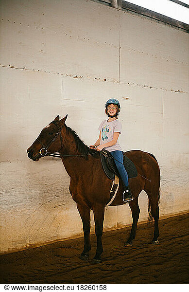 Happy girl on brown pony for horse back riding lesson in barn