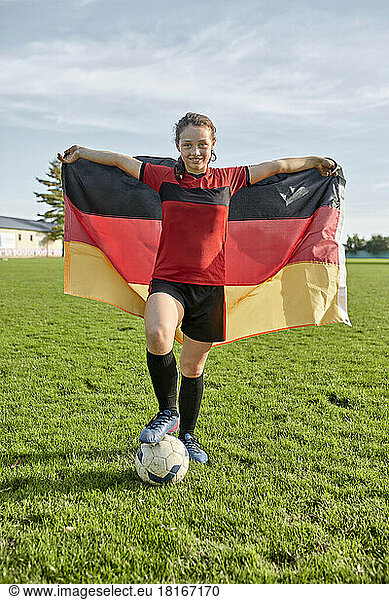 Happy girl standing with soccer ball and holding German flag on field