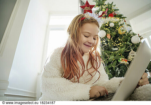 Happy girl using laptop sitting in front of Christmas tree at home
