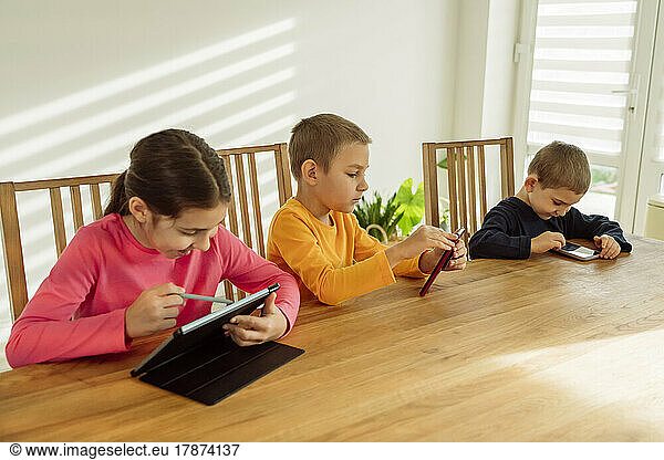 Happy girl using tablet PC sitting by brothers with technologies at home